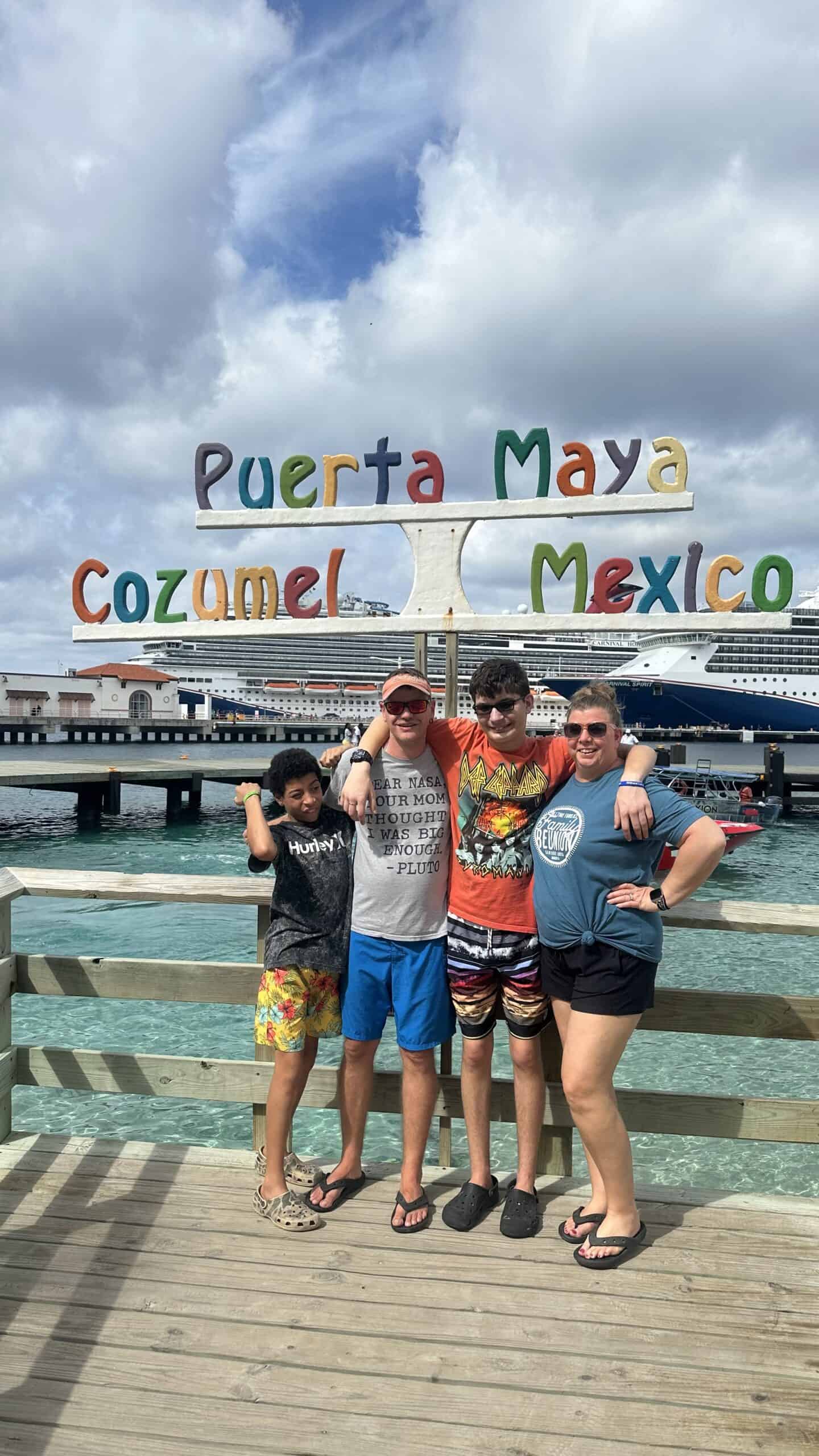 Four people standing in front of  a sign that  says Puerta Maya Cozumel Mexico. In the background is water and a cruise ship. 