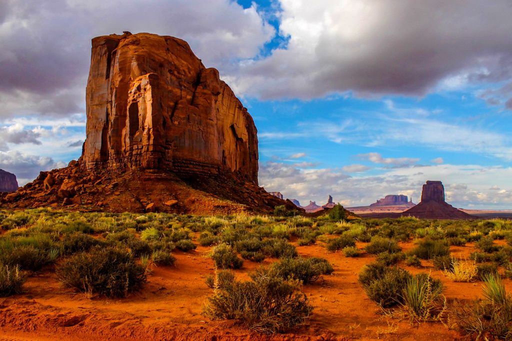 A stunning desert landscape featuring a large red rock formation under a partly cloudy sky. The foreground is covered with desert shrubs and red sand, while more rock formations stretch into the distance, capturing the vastness and beauty of the American Southwest.