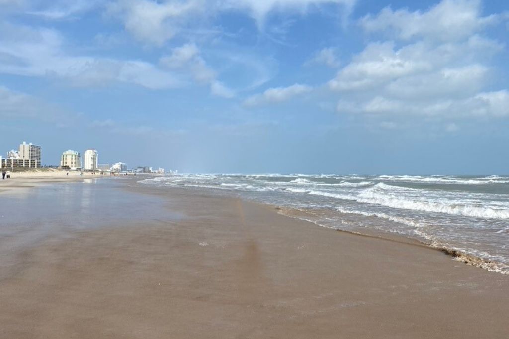 A wide sandy beach with gentle waves rolling onto the shore under a bright, partly cloudy sky. In the distance, several beachfront buildings line the coast, with a few people walking along the water’s edge, enjoying the peaceful, open space.