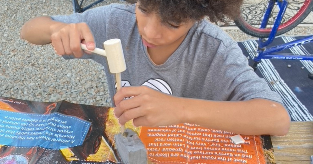 A child focused on an educational activity, using a small wooden hammer to chisel away at a geode stone on a work surface covered with an educational playmat. In the background, a bicycle is partially visible, suggesting the activity is taking place at a campsite.