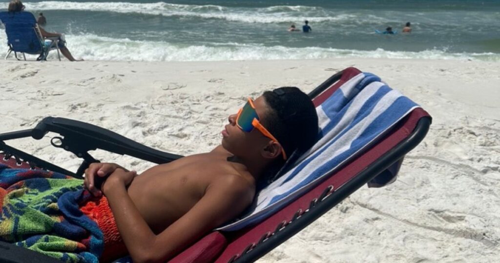 Tristan relaxing on a beach chair, sporting sunglasses, at Topsail Hill Preserve State Park in Florida, enjoying the white sandy beach and ocean waves with other beachgoers in the background.