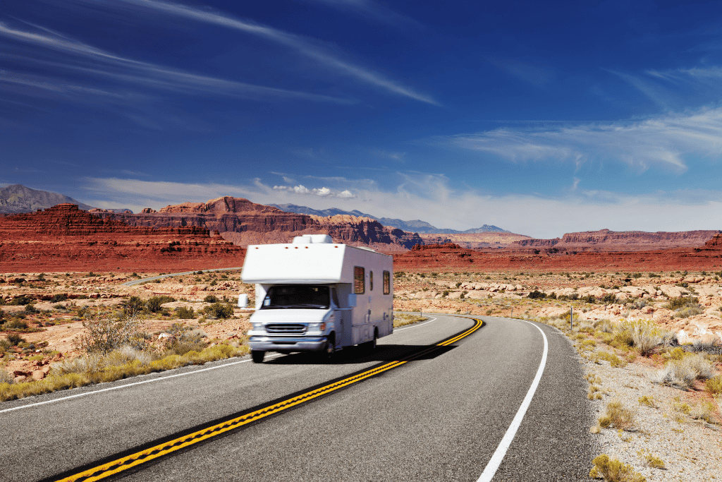 RV driving down the road with mountains in the background and desert all around. 