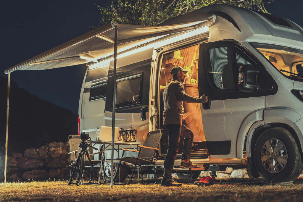 Man standing outside of a camper van peering in. There is an awning above him and chairs next to him. 