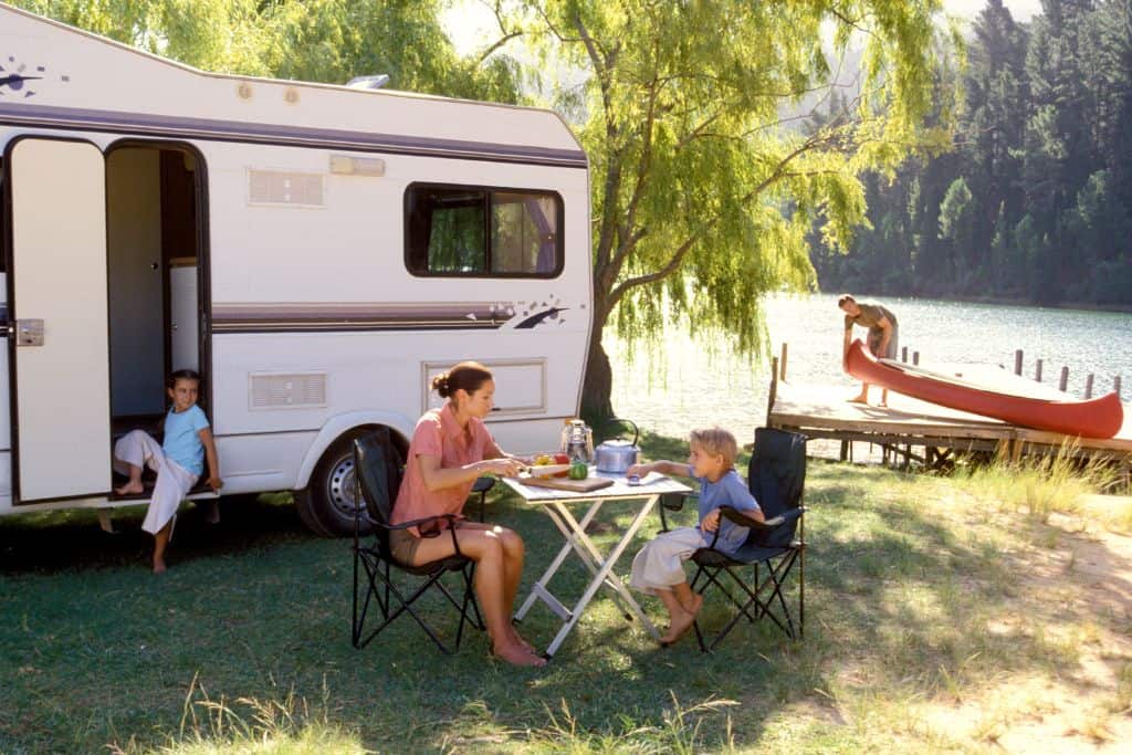 A serene camping scene by a lakeside with a family enjoying time outside their RV. A woman and a young boy are seated at a folding table, sharing a meal in camp chairs. Another child is sitting at the open door of the RV, and a man in the background is preparing to launch a red canoe from a wooden dock into the lake. The setting is lush with greenery, and the calm lake is surrounded by tall trees, creating a peaceful and relaxing atmosphere.