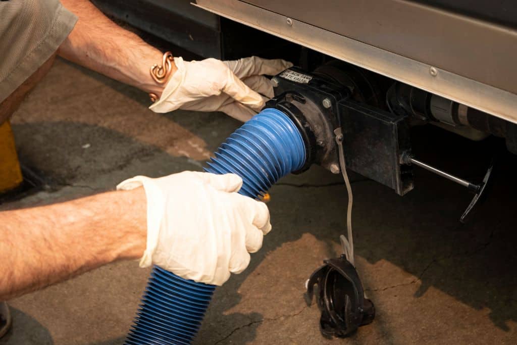 A close-up of a person wearing gloves connecting a blue sewer hose to the waste outlet of an RV. The process is part of emptying the RV's black and gray water tanks at a dump station. The person's hands are carefully securing the hose to ensure a proper connection, emphasizing the importance of sanitation and safety during the process.