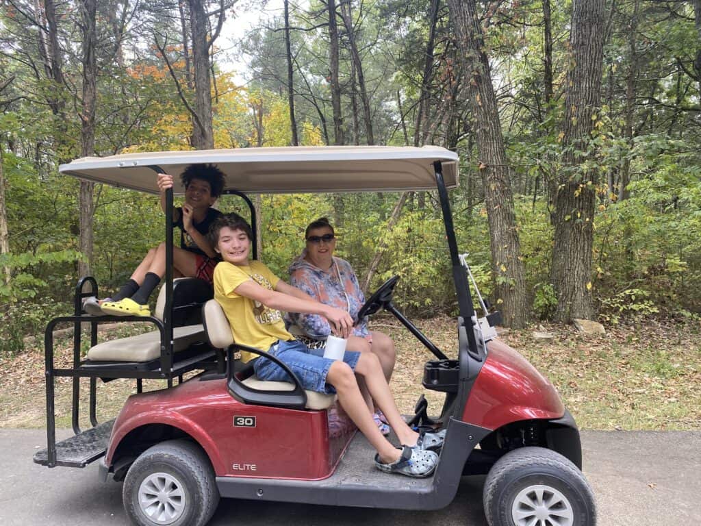 A group of three people riding in a red golf cart on a wooded path. Two boys, one sitting in the passenger seat and the other on the back seat, and an adult woman driving. The scene is surrounded by trees with green and yellow leaves, indicating early fall.