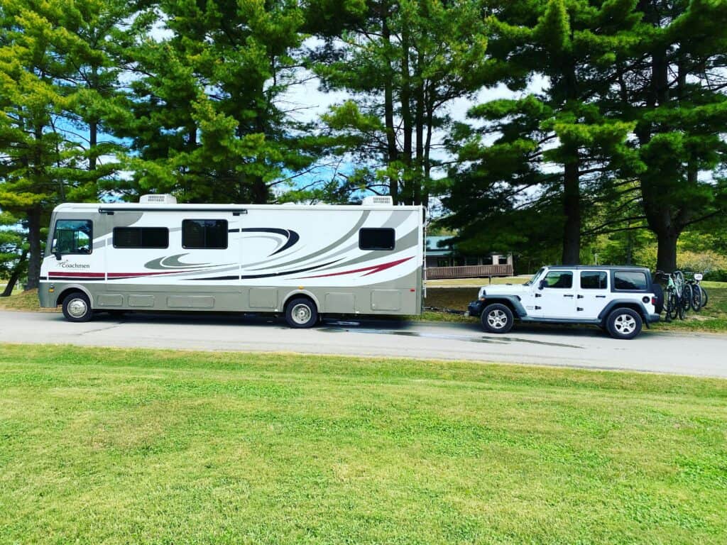 A large white motorhome with sleek gray and black accents, parked beside a row of tall pine trees. Attached behind the motorhome is a white Jeep with bikes mounted on the back. The setting is a sunny park with green grass and a clear sky.
