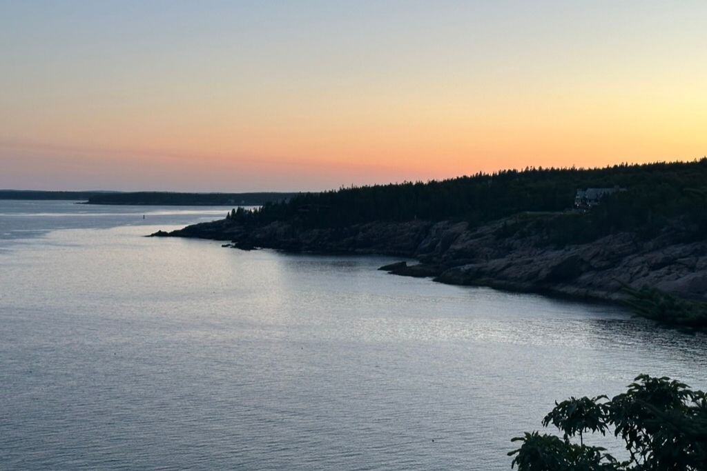 A serene coastal scene at sunset, featuring calm waters meeting rocky cliffs lined with trees. The sky fades from soft orange near the horizon to pale blue above, creating a peaceful twilight atmosphere. A small building is nestled among the trees on the cliffside, adding a sense of solitude to the landscape.