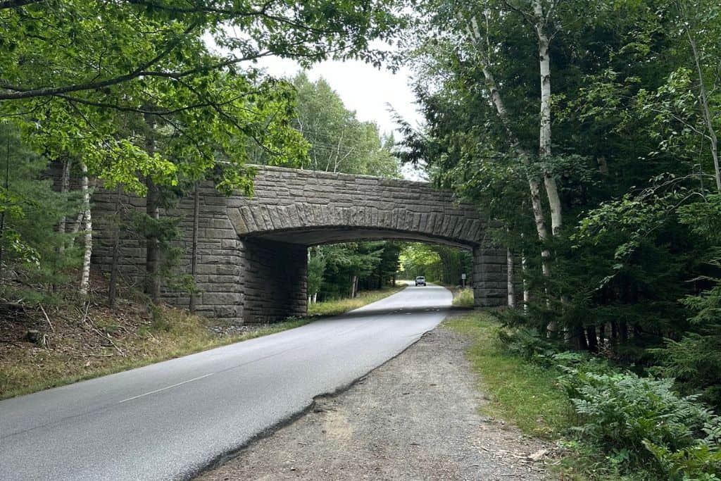 A stone bridge arching over a quiet, tree-lined road in a forested area. The bridge is made of large, rustic stone blocks, blending with the natural surroundings of tall birch and evergreen trees. A single car can be seen driving along the road under the bridge, adding to the peaceful, scenic atmosphere of this woodland setting.
