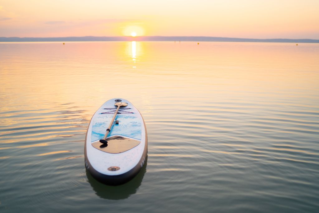 A serene scene of a paddleboard floating on calm water during sunset. The sky and water are bathed in soft pastel hues of orange, pink, and yellow, as the sun sets on the horizon, reflecting on the smooth surface of the lake. A paddle rests across the board, enhancing the tranquil and peaceful atmosphere.