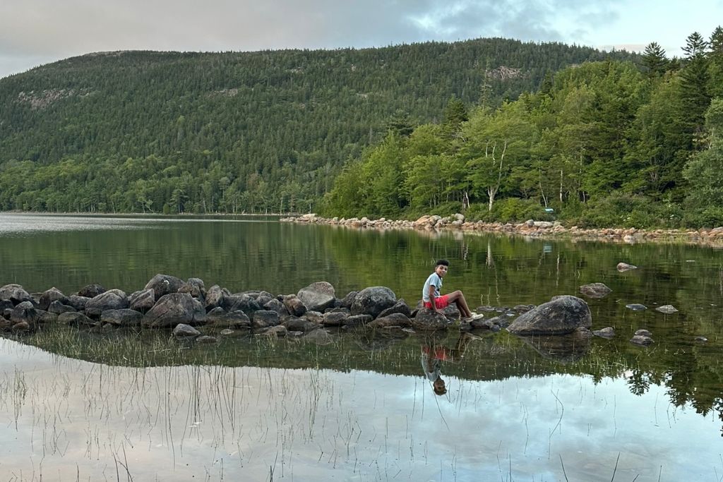 A young boy sits on large rocks in the shallow waters of a serene lake, surrounded by lush green forests and a forested mountain in the background. The still water reflects the trees, mountains, and the boy's image, creating a peaceful and symmetrical scene. The calm atmosphere and natural beauty of the area are emphasized by the tranquil setting.