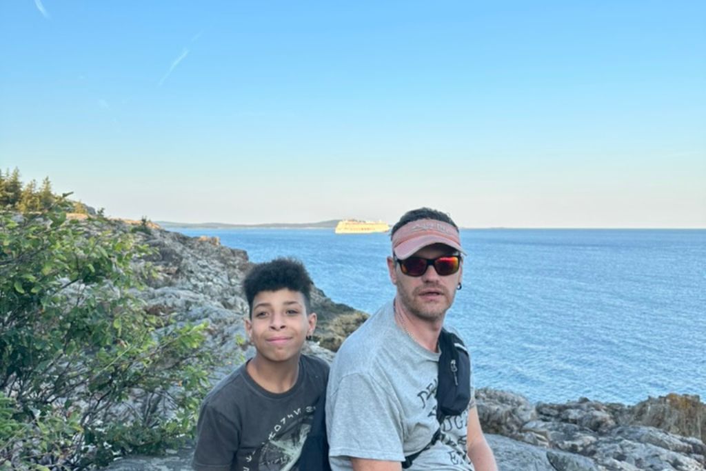 A man and a young boy sit together on rocky terrain near the edge of the ocean. Behind them, the deep blue water stretches out under a clear sky, with a distant cruise ship visible on the horizon. Both are casually dressed, with the man wearing sunglasses and a visor, while the boy smiles at the camera. The scene captures a moment of outdoor adventure and scenic beauty.