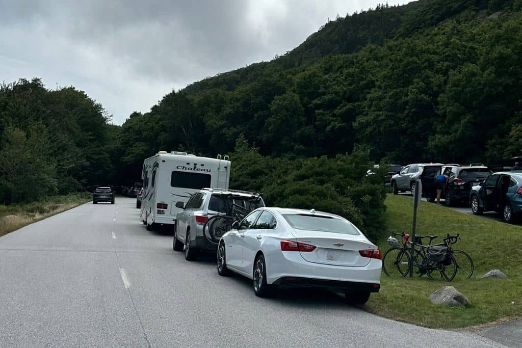 A row of vehicles, including an RV and cars with bike racks, parked along a scenic road surrounded by dense green trees and hills. The overcast sky casts a muted light on the scene, giving the area a calm, natural atmosphere. People can be seen standing near their vehicles, possibly preparing for outdoor activities, while bicycles are propped up against a signpost.