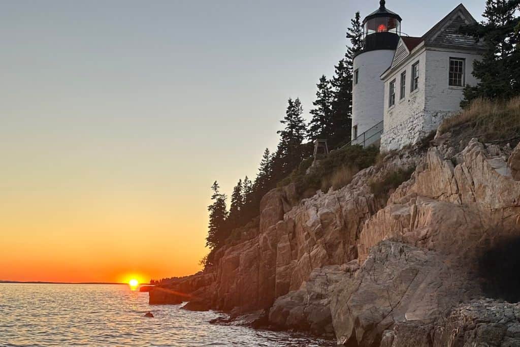 A breathtaking view of the Bass Harbor Head Lighthouse perched on rocky cliffs overlooking the ocean at sunset. The lighthouse's white exterior contrasts with the orange and pink hues of the sky as the sun sets on the horizon, casting a warm glow on the water and rocks below. Tall pine trees surround the lighthouse, adding to the serene and picturesque coastal scene.