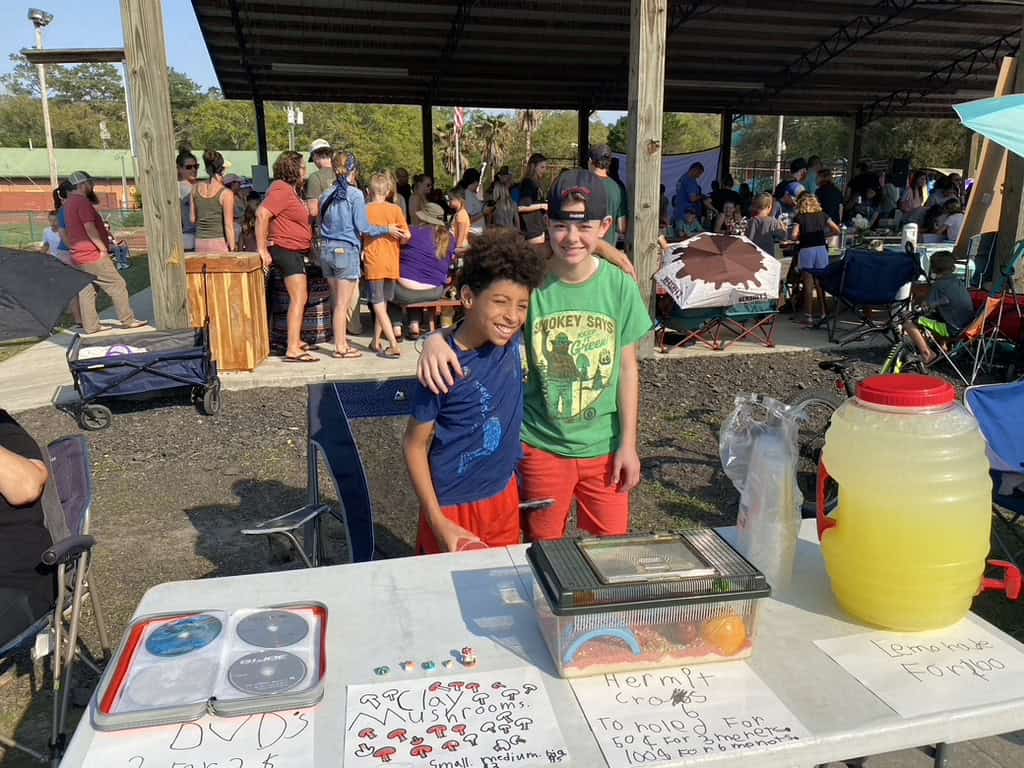 Two kids with arms around each other behind a table with a jar of lemonade, a hermit crab cage and DVD's. In the background there are people gathering under a pavillion.