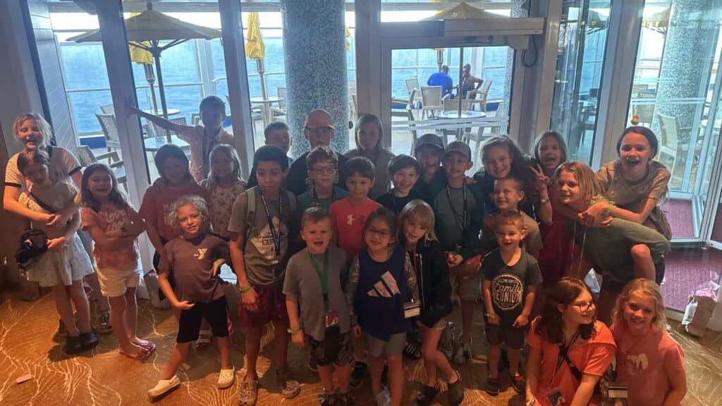 Large group of kids standing in front of a glass wall. In the background there are people sitting at a table.