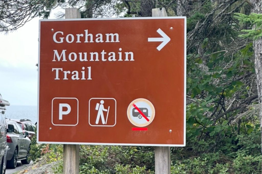 A brown trailhead sign indicating the direction to the 'Gorham Mountain Trail' with an arrow pointing to the right. Below the trail name are symbols for parking, hiking, and a prohibition on trailers or RVs. The sign is situated near trees, with parked cars visible to the side, and likely marks the beginning of a hiking trail in a scenic area.