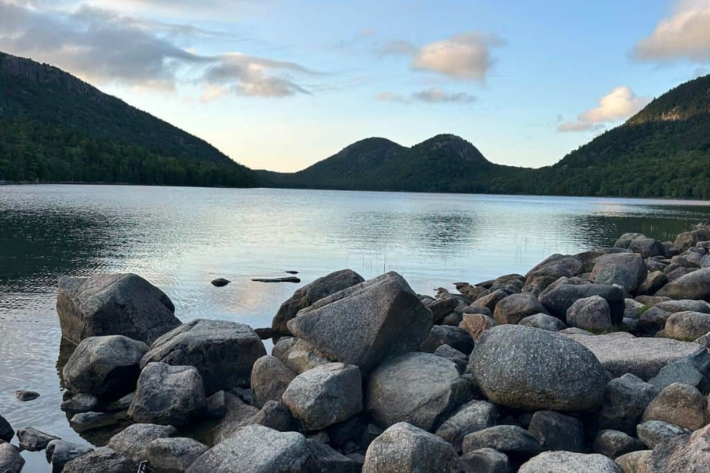 This image shows a peaceful view of Jordan Pond in Acadia National Park, with rocky shores in the foreground and calm water reflecting the sky. In the background, two rounded hills known as the Bubbles stand prominently, surrounded by lush green forests. The scene captures the serene beauty of the park in soft, natural light, with scattered clouds in the sky above.