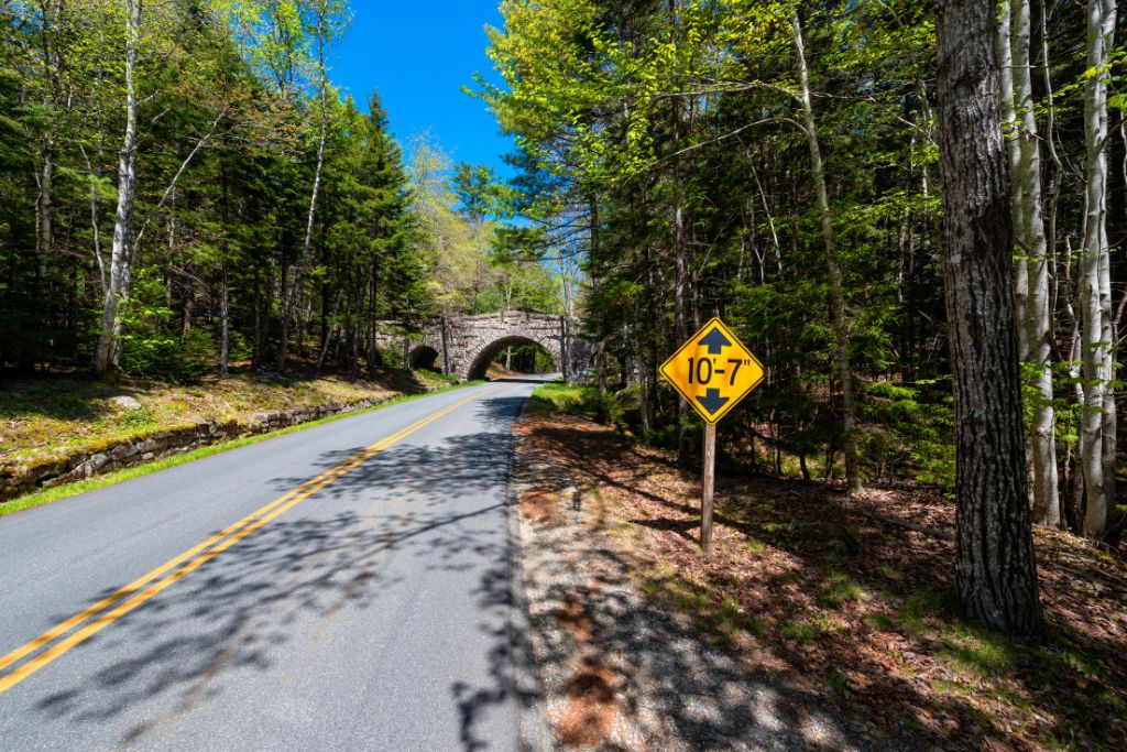 This image shows a scenic stretch of road in Acadia National Park, bordered by tall, leafy trees. In the distance, a stone bridge arches over the road, framed by greenery and illuminated by bright sunlight. A yellow height clearance sign (10'-7") is posted by the roadside, warning drivers of the low bridge. The peaceful forest setting and clear blue sky add to the tranquil atmosphere of this well-known Park Loop Road.