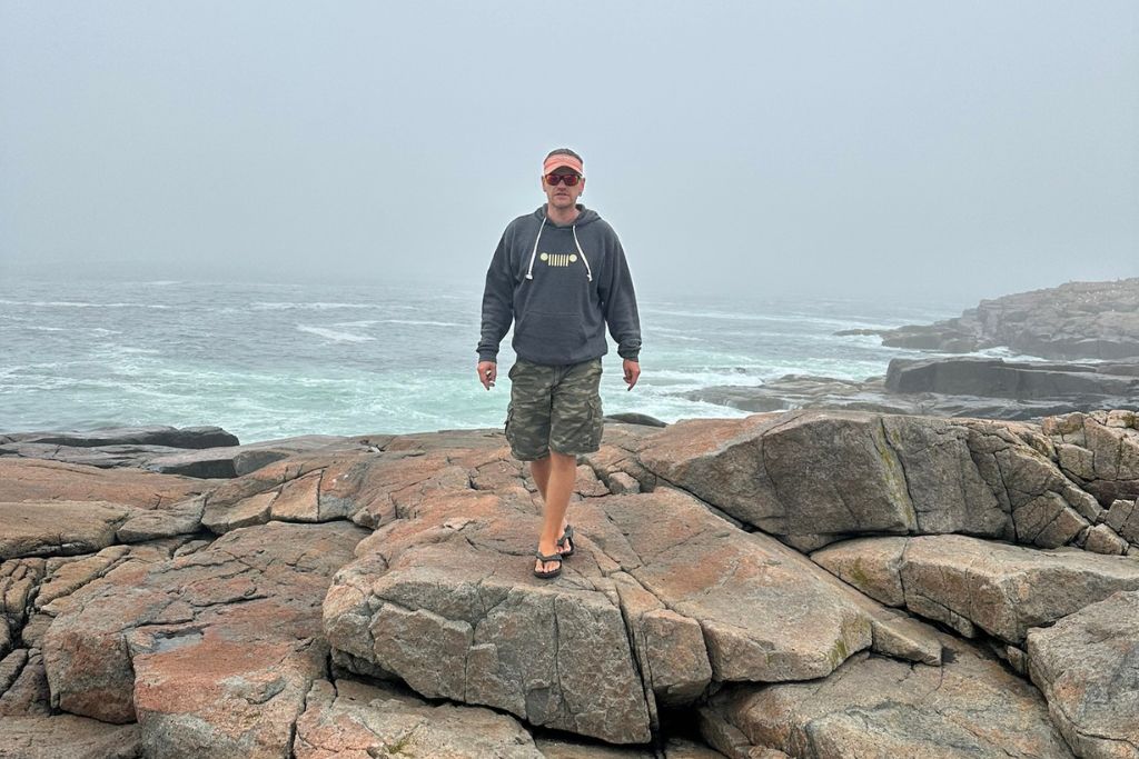 This image shows a person standing on large, rugged rocks along the coast of Acadia National Park. The ocean waves crash against the rocky shoreline in the background, with a foggy sky creating a misty and dramatic atmosphere. The person, dressed in a hoodie, camouflage shorts, and flip-flops, appears to be enjoying the coastal scenery despite the overcast weather. The scene captures the raw beauty of Acadia’s coastline, even on a misty day.