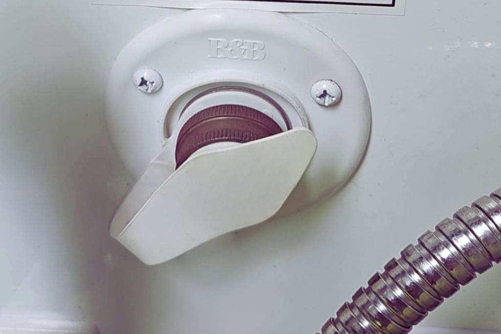 A close-up of an RV water fill inlet with a white twist-lock cap, marked with the brand "B&B." The cap is partially open, allowing access for filling the fresh water tank. A flexible metal hose is visible nearby, suggesting a connection for water supply.
