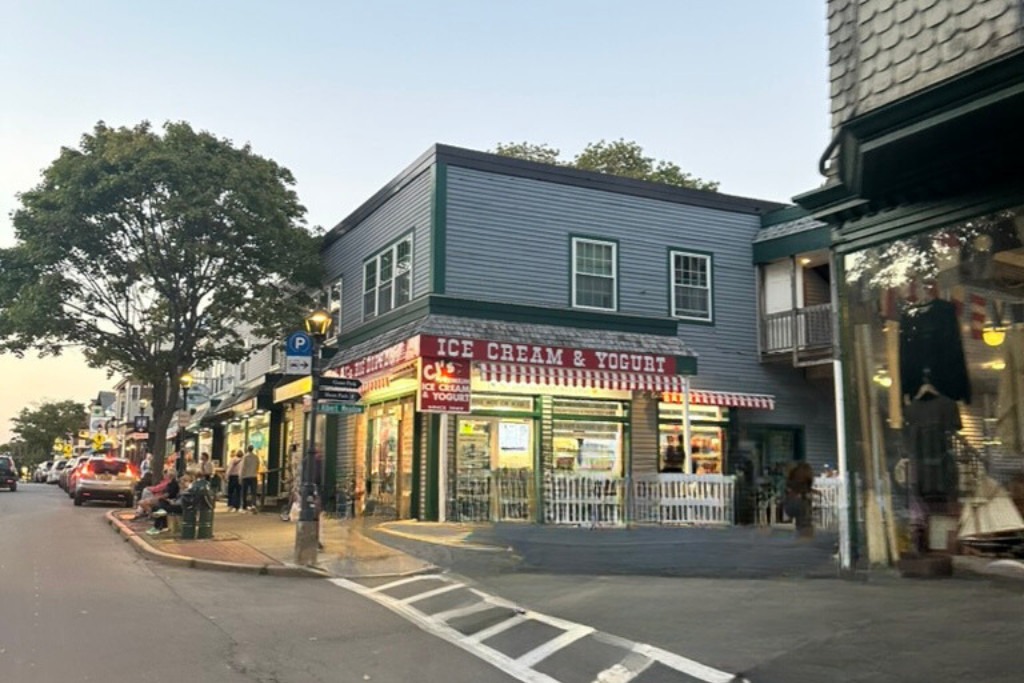 A bustling street scene at dusk featuring an ice cream and yogurt shop with a red and white striped awning. People are gathered on the sidewalk, and cars are parked along the street. Trees line the area, and nearby buildings showcase small-town charm with quaint storefronts.