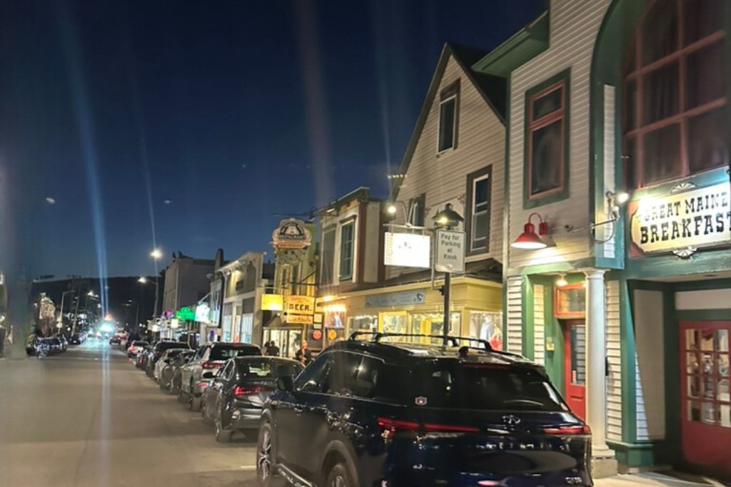 A lively street at night with cars parked along the curb and brightly lit storefronts, including a sign for "Great Maine Breakfast." The sky is dark, and the street is illuminated by streetlights and neon signs, creating a cozy small-town evening atmosphere.