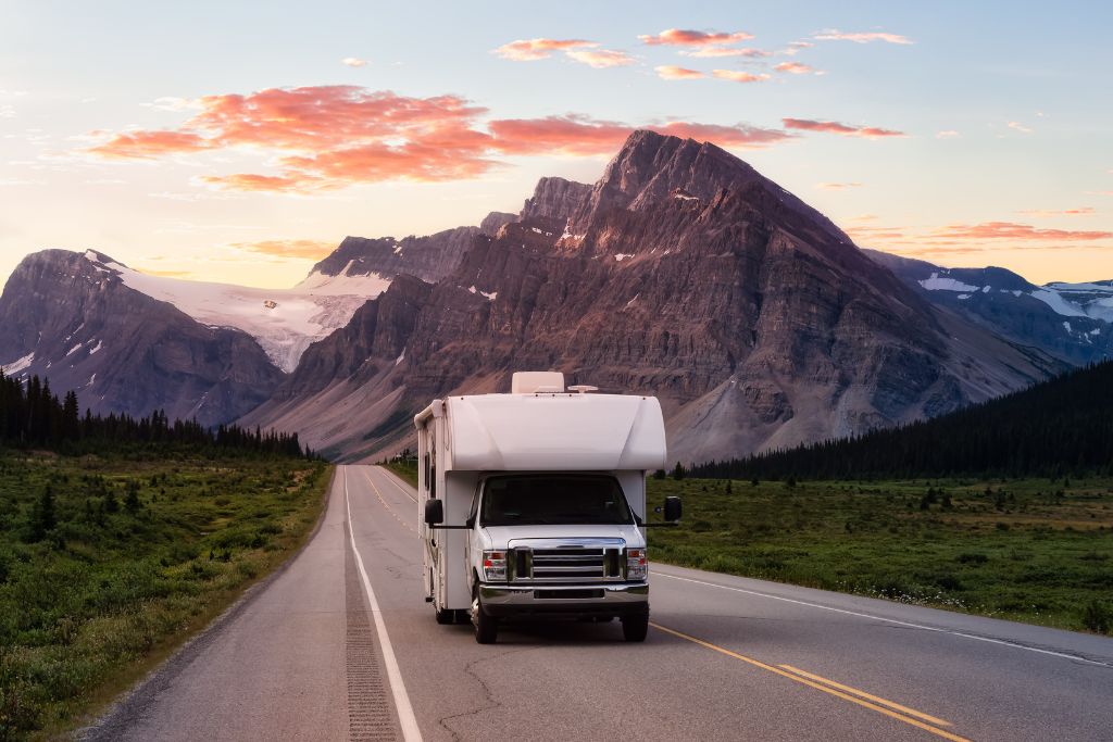 An RV travels on a scenic mountain road at sunset, surrounded by towering peaks and lush green landscape. The sky is painted with soft pink and orange clouds, casting a warm glow over the mountainous terrain.