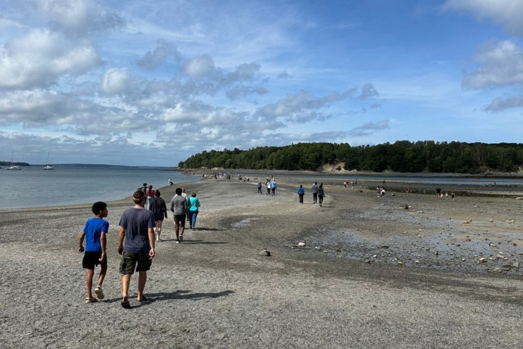 A group of people walking along a rocky sandbar that stretches out into the ocean under a partly cloudy sky. The sandbar connects to a tree-covered island in the distance, with boats anchored nearby.