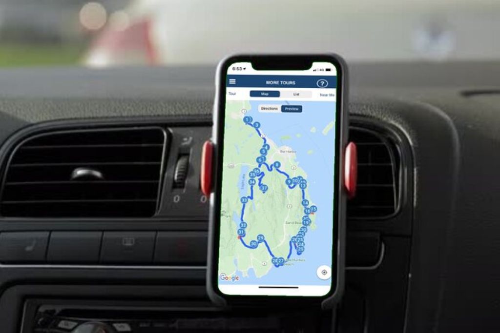 This image shows a smartphone mounted on a car dashboard, displaying a map with a detailed route for a driving tour around Acadia National Park. The screen highlights numerous stops along the route, guiding visitors through various points of interest in the park. The setup offers a convenient, hands-free way to explore Acadia’s attractions, allowing for a self-guided tour experience with navigation and information readily accessible on the phone.