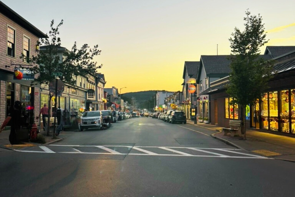 A quaint small-town street at sunset with soft golden light. Lined with parked cars, storefronts, and glowing signs, the street exudes a peaceful evening atmosphere. A few pedestrians walk along the sidewalks, and a distant hill is visible at the end of the street.