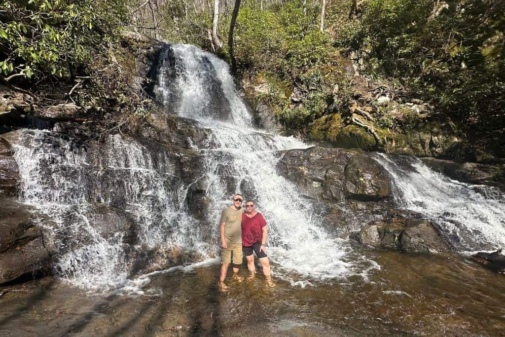 The image shows two people standing in front of a stunning waterfall cascading over dark rocks. They are smiling and standing ankle-deep in clear water at the base of the falls. The area is surrounded by lush greenery, with moss-covered rocks and trees framing the waterfall. The bright sunlight highlights the water's movement, creating a vibrant and refreshing scene.