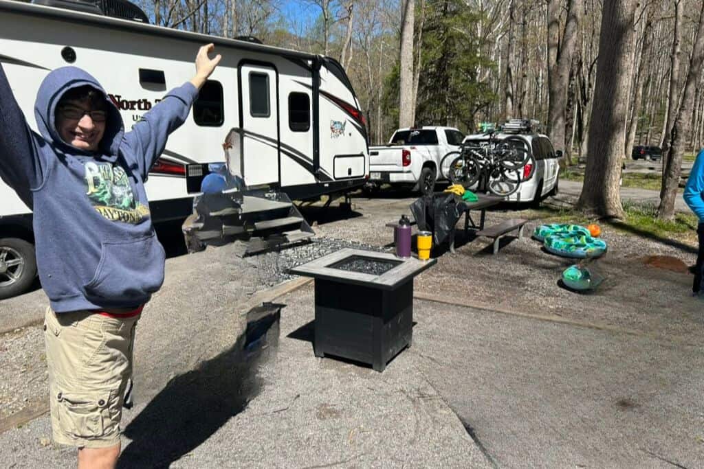 The image shows a lively campsite with a cheerful person in the foreground, raising their arms and smiling. Behind them, there’s a white camper trailer parked alongside a truck and SUV. The site is equipped with a picnic table holding supplies like water bottles and a bike rack loaded with bicycles. Camping gear, including pool floats, is scattered nearby, and a portable fire pit is set up in the foreground. The area is surrounded by tall trees, suggesting a woodland setting on a bright, sunny day.