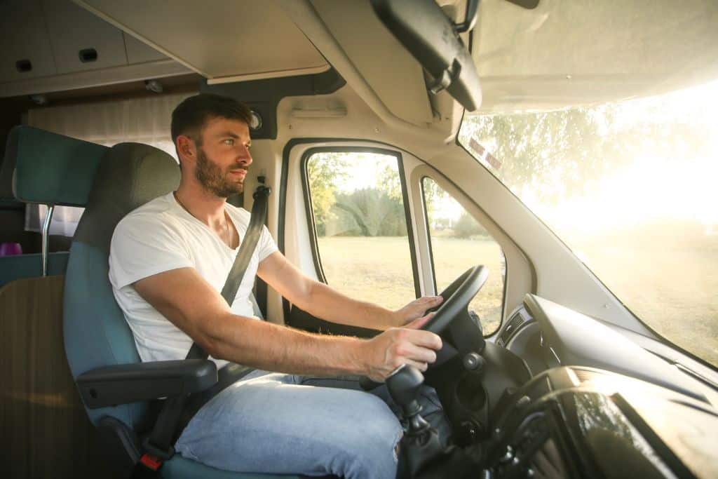 A man with short brown hair and a beard is seated behind the wheel of an RV, driving during golden hour. He wears a white t-shirt and a seatbelt, with sunlight streaming through the windshield, creating a warm glow. The background reveals a glimpse of trees and a grassy area outside.