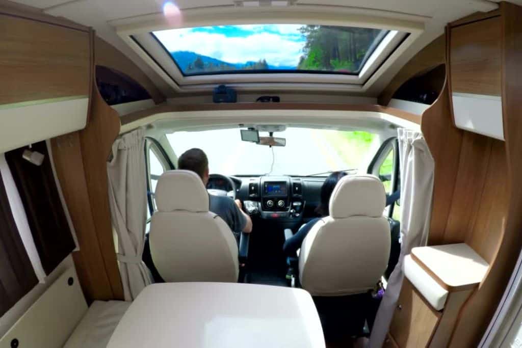 This is an interior view of an RV, showing two people seated in the front cab—one driving and the other in the passenger seat. The perspective is from the back of the vehicle, highlighting the modern, cozy wood-paneled interior and a large skylight above. The road ahead is visible through the windshield, surrounded by greenery and a clear blue sky seen through the skylight, creating a scenic and inviting atmosphere.