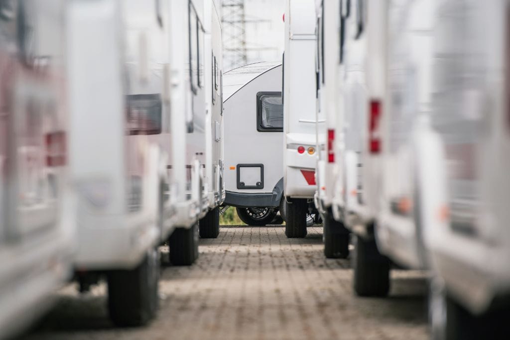 
This image shows a row of parked white RVs or camper trailers on either side, focusing on a single RV at the center in the background. The perspective emphasizes the symmetry of the parked vehicles on a paved surface, creating a clean and organized composition.