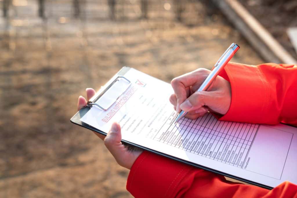 This image shows a person in a bright orange jacket holding a clipboard with a "Building Inspection Report" form, partially filled out with a pen. The background appears to be a construction site with blurred earthy tones, emphasizing the inspection context.