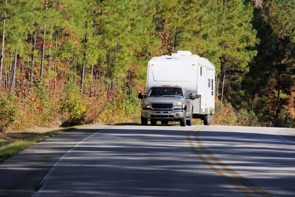 This image shows a pickup truck towing a white RV on a curving road surrounded by lush greenery and autumn-colored trees. The bright, natural setting emphasizes a peaceful journey through a scenic route, ideal for road trips or camping adventures.