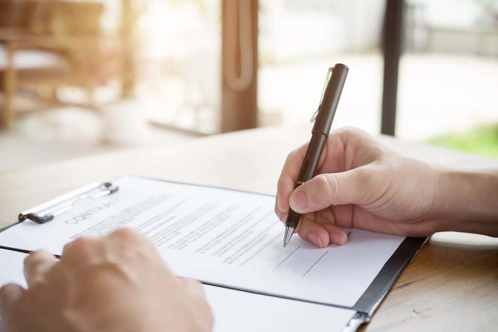 This image depicts a person signing a document labeled "Contract" on a clipboard placed on a wooden table. The bright, softly lit background suggests a calm indoor setting, highlighting the act of agreement or formalizing a deal.